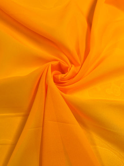 A close-up view of the vibrant Saffron Chiffon from Super Cheap Fabrics, showcasing its lightweight and smooth-textured fabric. The material is delicately draped in a circular, swirling pattern at its center, creating soft folds and gentle shadows. The bright, vivid orange radiates warmth and vibrancy, emphasizing the fluidity and elegance of this 150cm wide fabric.