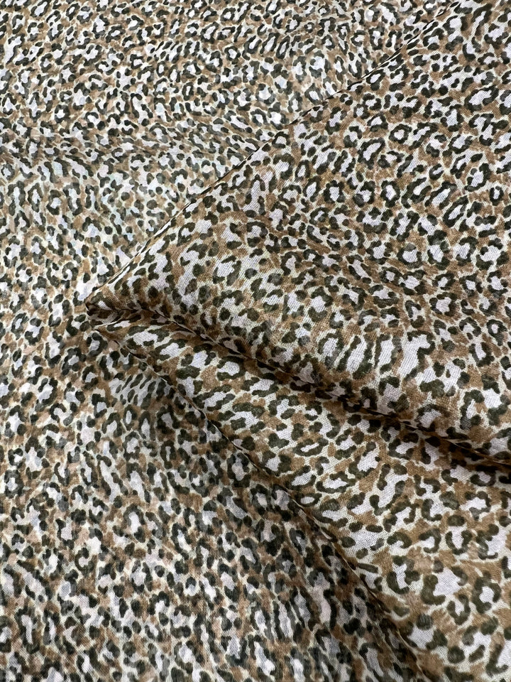 A detailed image featuring the Pure Printed Silk - Namir from Super Cheap Fabrics, showcasing a leopard print pattern. The multi-colour design includes brown, black, and white spots for a textured and natural appearance. Some sections of the 75 GSM fabric are folded to highlight its flexibility and softness. The fabric measures 135cm in width.