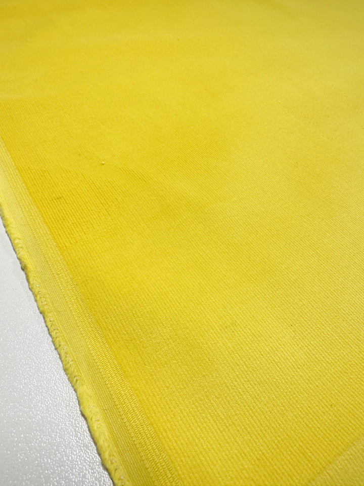 Close-up of Micro Wale Corduroy - Golden Kiwi by Super Cheap Fabrics draped over a white surface. The ribbed texture is slightly textured, with a neatly sewn edge visible along the left side, creating sharp contrast with the white background. Fabric width: 142cm.