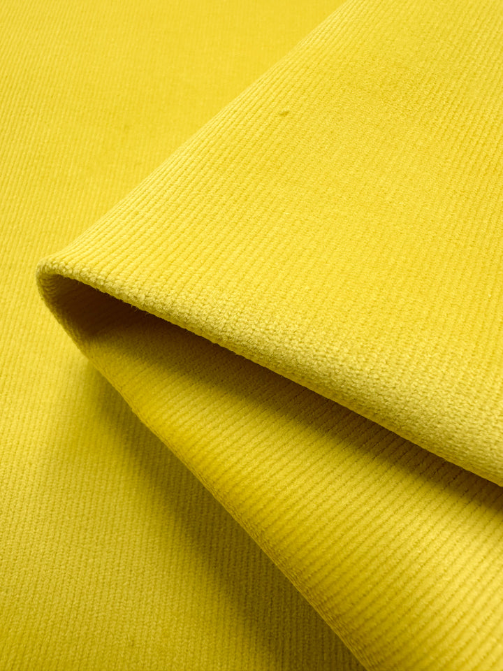 The Micro Wale Corduroy - Golden Kiwi by Super Cheap Fabrics features a textured yellow appearance, ideal for winter clothing. Its layered creases and shadows highlight the thick diagonal ribbed texture, adding depth to the vibrant 142cm-wide material.