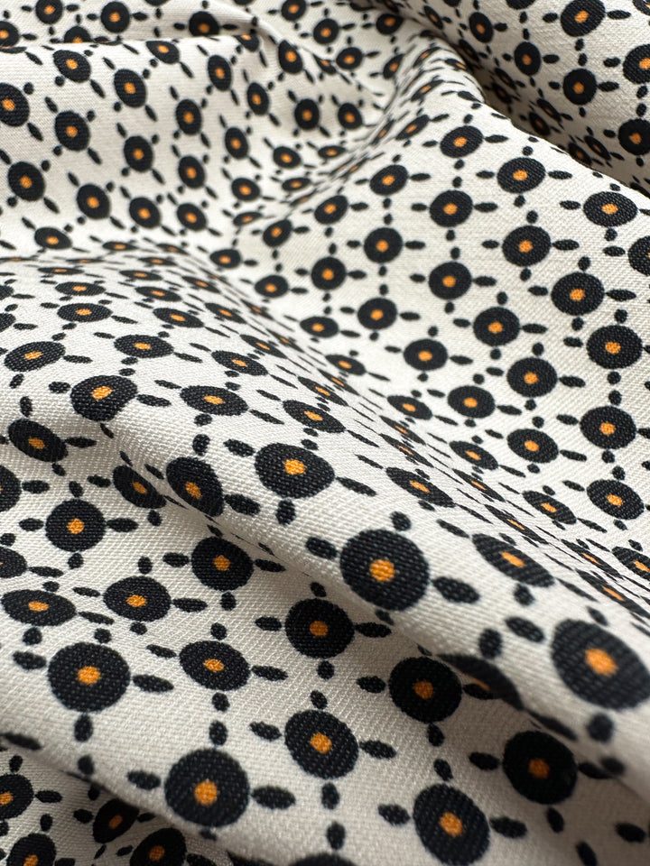 A close-up of Super Cheap Fabrics' Printed Bengaline - Radar - 150cm reveals a medium-weight fabric with a black and orange geometric floral pattern on beige. The slightly wrinkled surface highlights the texture and intricate design of this two-way stretch woven print.