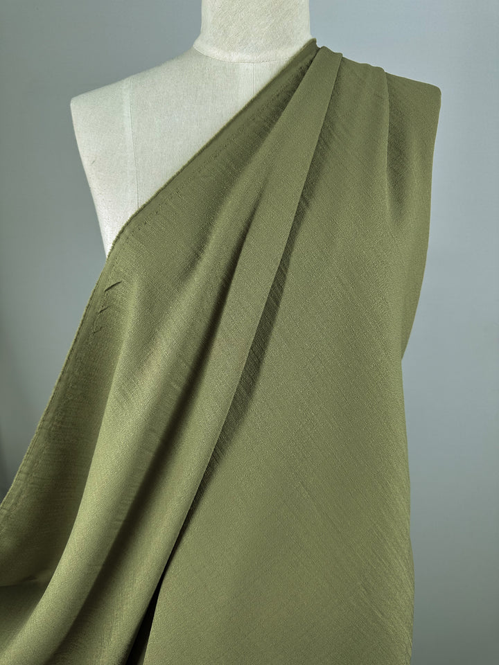 A mannequin displays the texture and flow of Super Cheap Fabrics' Textured Suiting in Cedar, an olive green, lightweight fabric draped asymmetrically over the shoulder against a neutral background.