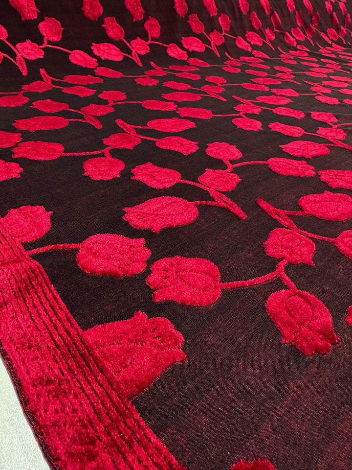 The Burnout Velvet - Lychee by Super Cheap Fabrics features luxurious fabric with intricate red, textured tulip designs. The vibrant contrast between the red flowers and dark background creates an elegant pattern that uniformly covers the 150cm wide fabric.