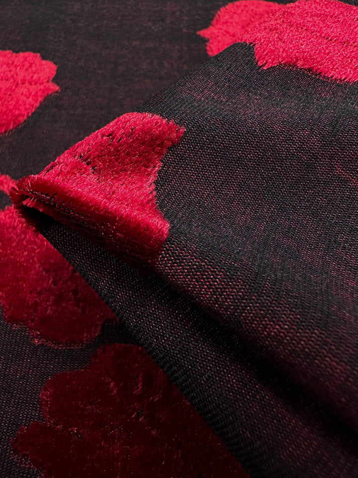 Close-up of Burnout Velvet - Lychee by Super Cheap Fabrics, featuring luxurious black fabric with vibrant red floral designs. The intricate pattern on the textured surface highlights rich colors, evoking an elegant contrast against the dark background.