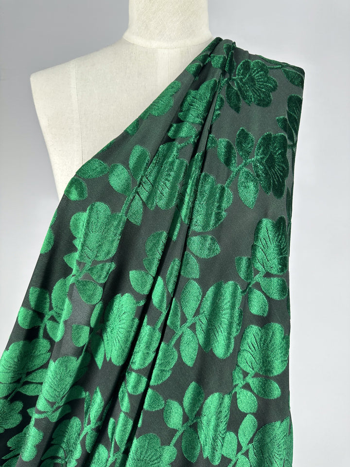 A luxurious Burnout Velvet - Amazon - 150cm from Super Cheap Fabrics elegantly drapes over a mannequin, featuring a dark green base with an intricate pattern of large, glossy green leaves, exuding sophistication and elegance with its rich texture.