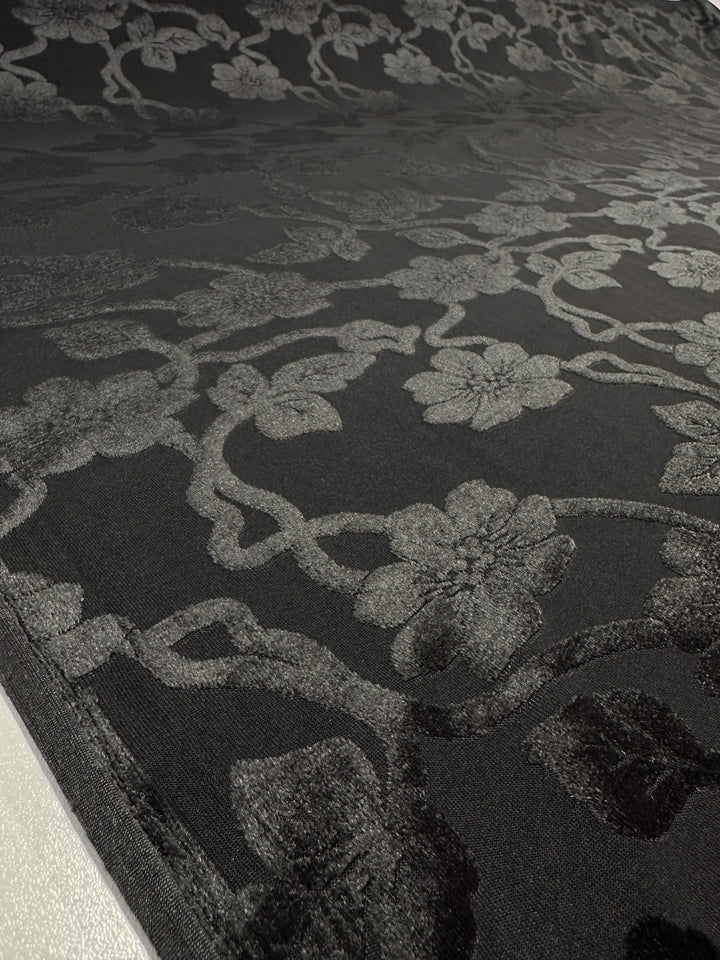 A close-up of the Super Cheap Fabrics Burnout Velvet - Noir (130cm) reveals an intricate floral pattern in gray. The luxurious design, with vines and flowers, creates a textured elegance, highlighted beautifully by the lighting.