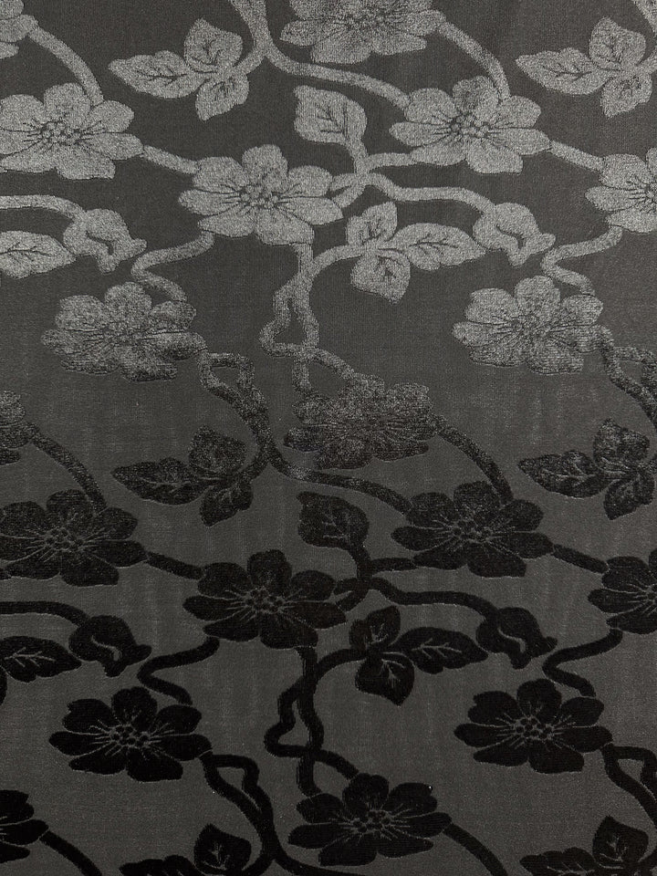 The Burnout Velvet - Noir - 130cm by Super Cheap Fabrics features an intricate floral pattern with a luxurious gradient of flowers transitioning from light gray to dark black on a muted gray background, showcasing elegantly intertwined stems and leaves.