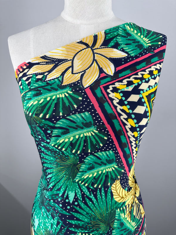 A mannequin displays the vibrant, medium-weight Super Cheap Fabrics' Palm Springs Printed Lycra, featuring a one-shoulder style. The fabric showcases bold patterns with large yellow flowers, lush green leaves, and pink/yellow/black geometric details for a tropical theme.