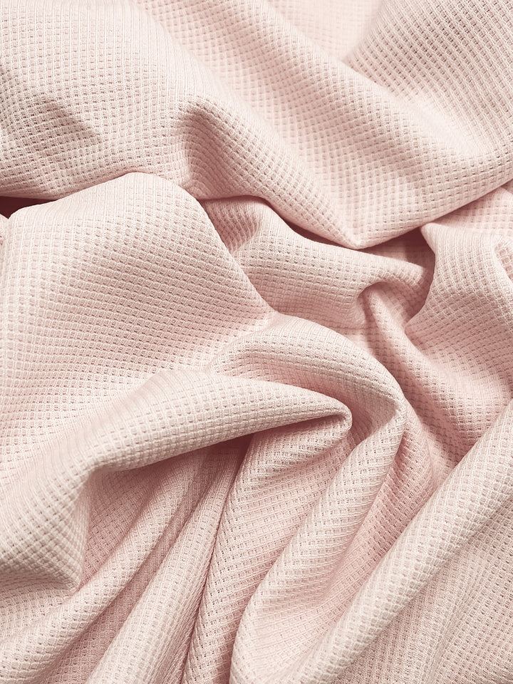 Close-up of the soft, pearl blush Waffle Knit fabric by Super Cheap Fabrics, featuring a textured honeycomb design with a waffle weave pattern. The material is gently draped, creating folds and shadows that enhance its three-dimensional appearance.