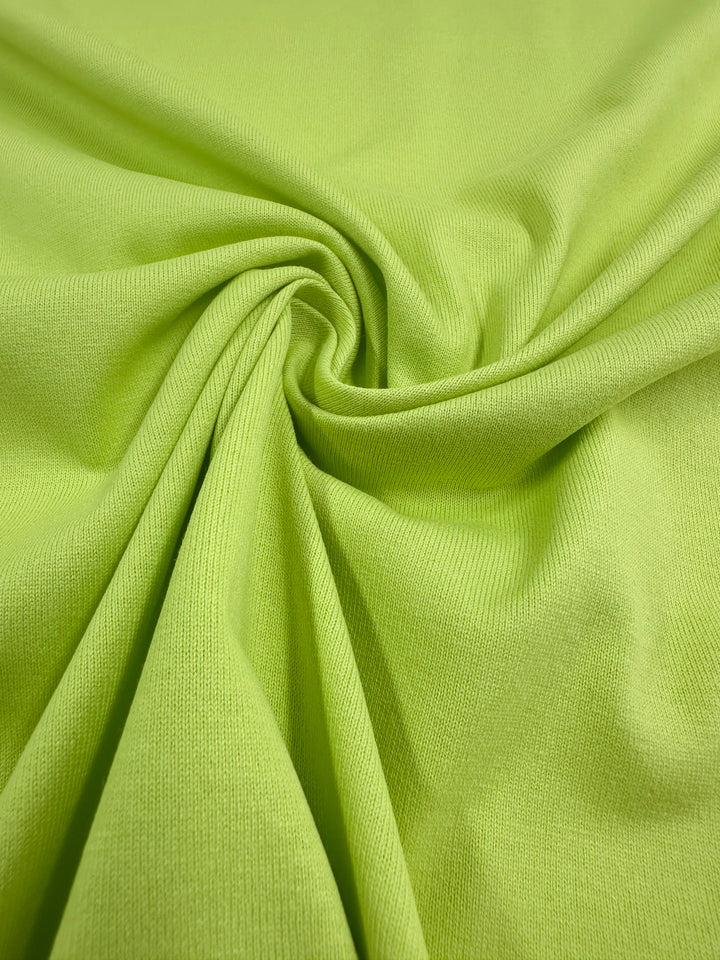 A close-up of Super Cheap Fabrics' Daiquiri Green Cotton Jersey showcases folds and creases in a centered swirl. The smooth, even weave promises the comfort typical of jersey cotton, making the 170cm textile truly inviting.