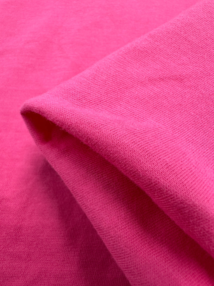 A close-up of Super Cheap Fabrics' Cotton Jersey - Fandango Pink, 160cm wide, reveals its vibrant color and smooth texture. The image features a fold that highlights the material's softness and weave.