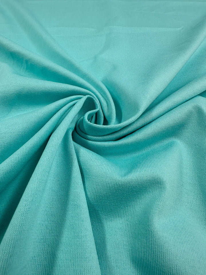 A close-up of Super Cheap Fabrics' Cotton Jersey - Aqua Splash - 160cm shows a textured woven pattern with the cotton gathered in a circular fold at the center, creating a visual swirl.