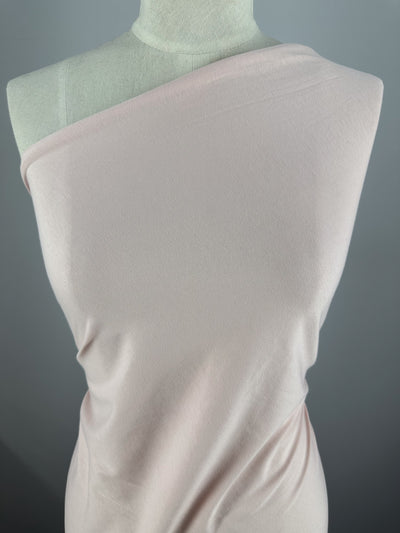 A light pink, one-shoulder dress in Rosewater made from Super Cheap Fabrics' Cotton Jersey (165cm) is elegantly draped on a mannequin against a gray background. This versatile piece boasts soft cotton fabric with a gentle sheen.