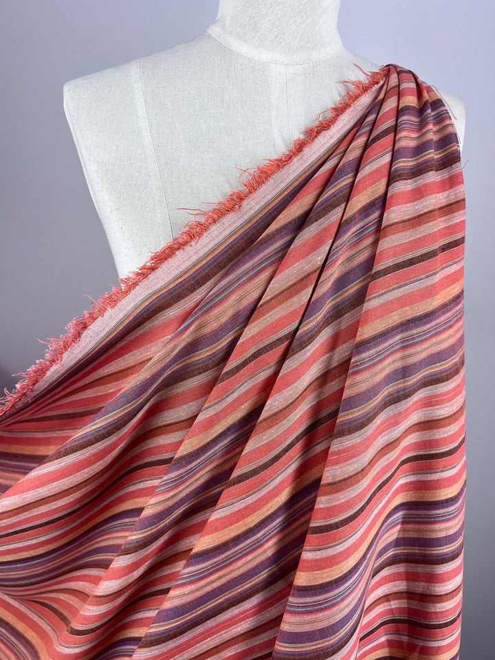 The Linen Blend - Colombo - 150cm fabric from Super Cheap Fabrics, featuring a yarn-dyed, striped design in shades of pink, purple, and orange, elegantly drapes over a mannequin. Its frayed edges and diagonal lines contribute to its vintage appearance with a colorful pattern against a plain white-gray background.