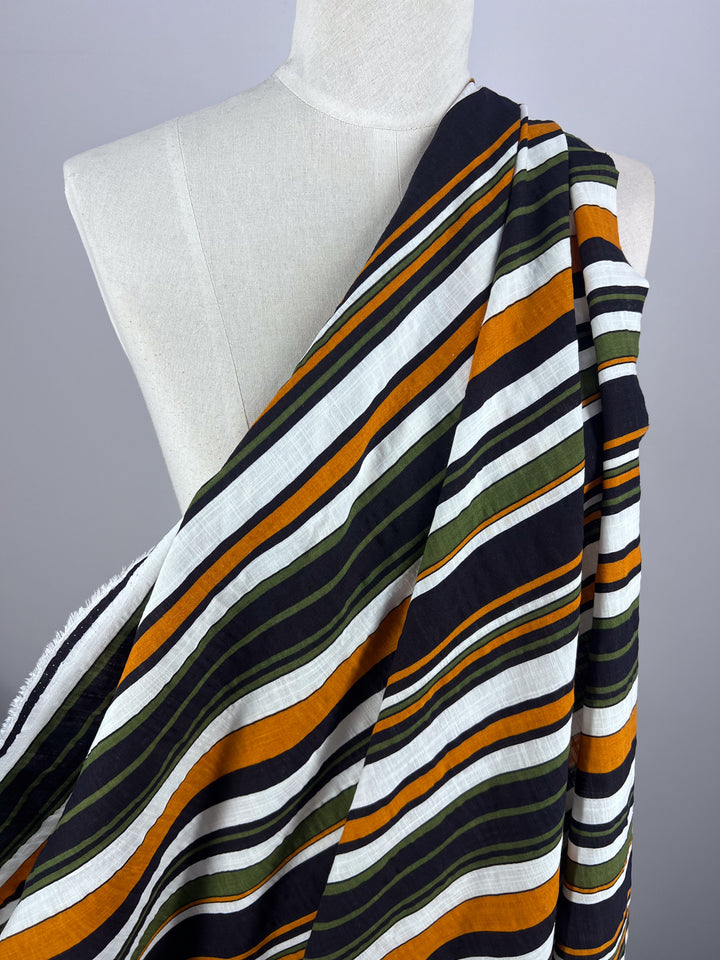 A mannequin displays the Linen Blend - Retro - 145cm from Super Cheap Fabrics, featuring breathable material perfect for summer attire, effortlessly draped over one shoulder. The fabric is adorned with bold horizontal stripes in black, orange, green, and white.