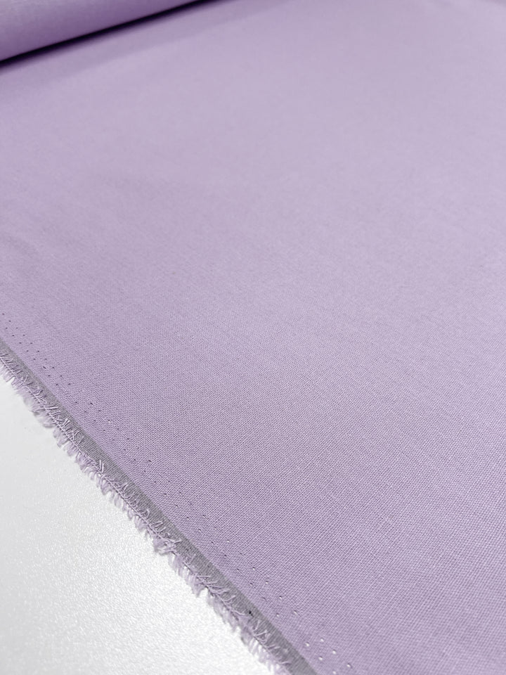 Close-up of a lavender-colored fabric from Super Cheap Fabrics, known as the Premium Linen Cotton - Orchid Bouquet - 140cm, showcasing its textured edge. Laid flat on a white surface, this lightweight material exhibits a smooth and soft appearance, emphasizing its fine weave and subtle sheen reminiscent of an elegant orchid bouquet.