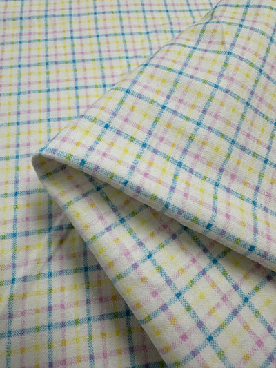 A close-up image of a folded piece of fabric showcasing a plaid pattern. This 100% cotton, lightweight fabric has a white background with intersecting lines in blue, pink, yellow, and green, forming a multi-colour checkered design. The texture appears soft and slightly fuzzy. The product is Flannelette - Tattersall - 110cm by Super Cheap Fabrics.