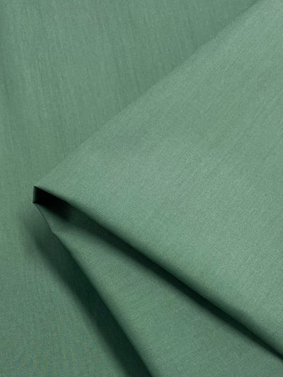 A close-up of lightweight, folded Loden Frost poplin from Super Cheap Fabrics highlights its smooth texture and even coloring. Neatly arranged with clean lines, the fabric captures a subtle play of light and shadow—ideal for crafting durable childrenswear. Measures 148cm wide.