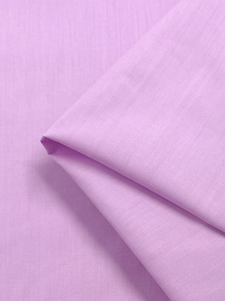 A close-up of the Poplin - Pastel Lavender fabric by Super Cheap Fabrics, folded neatly. This 148cm polyester-cotton blend is lightweight with a smooth texture and subtle sheen, its delicate hue highlighted by soft lighting.