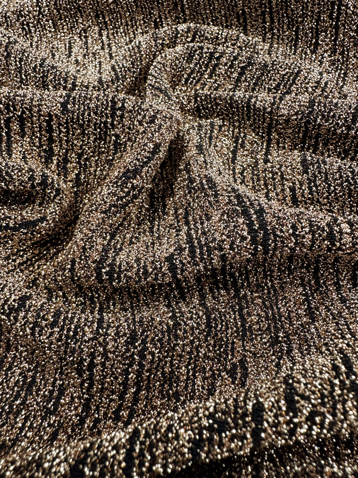 Close-up of Super Cheap Fabrics' Metallic Lycra - Golden, 150cm wide, showcasing a shimmering gold and black textured pattern. The surface catches the light, highlighting its metallic sheen and intricate design—ideal for crafting elegant dresses or skirts.