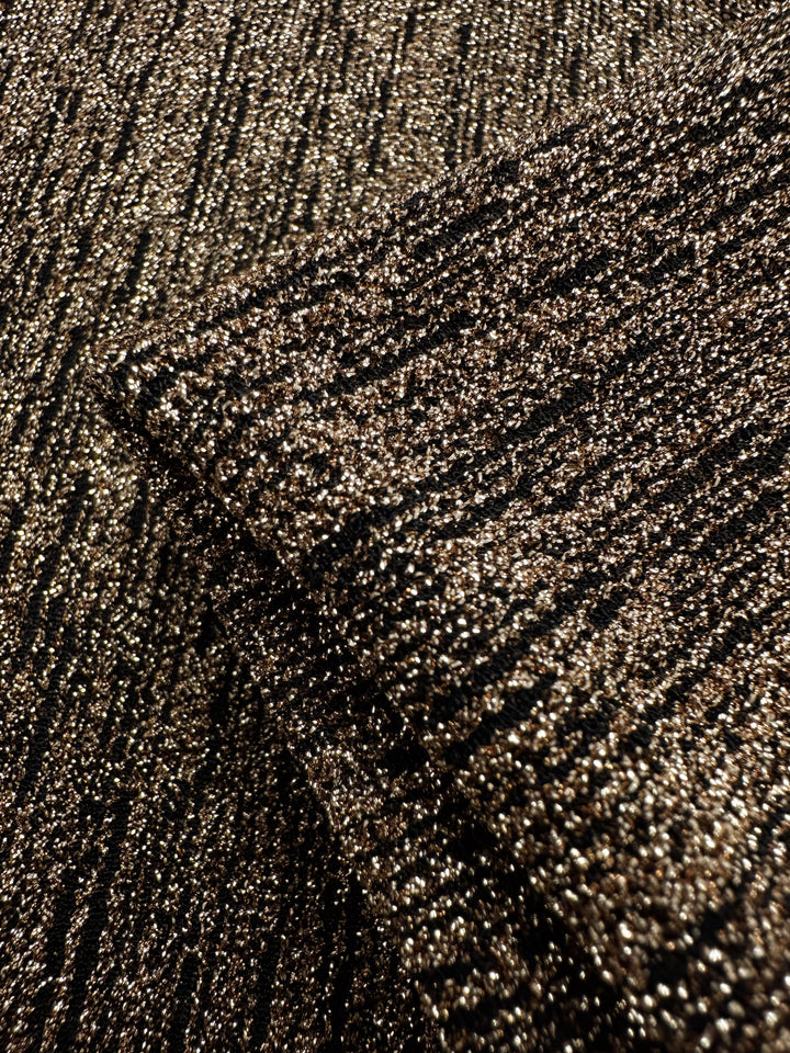 A close-up of Super Cheap Fabrics' Metallic Lycra - Golden - 150cm shows a textured fabric with shimmering gold and copper threads, giving it a glittery effect. The medium-weight lycra is folded, highlighting its intricate pattern and reflective quality, perfect for dresses and skirts.