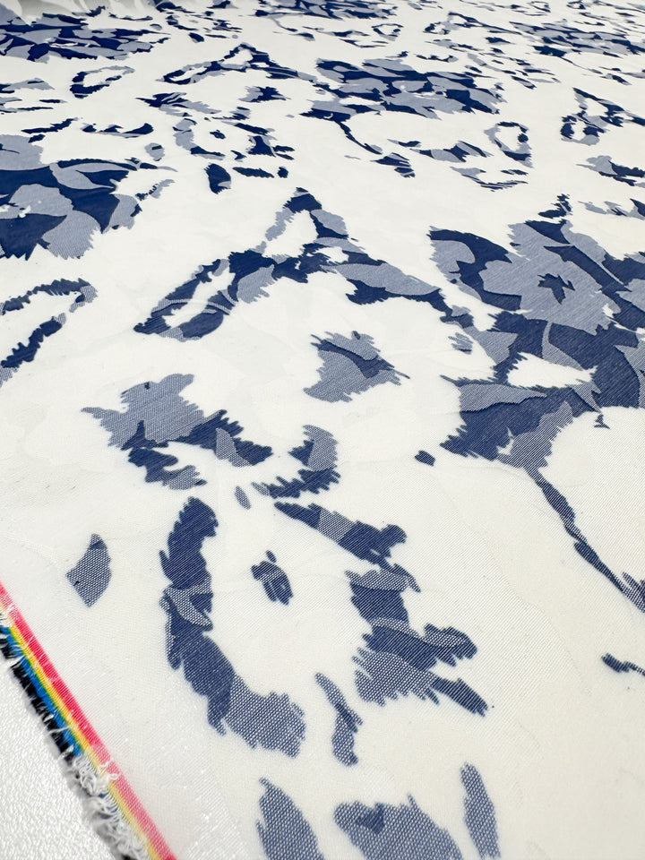 The Deluxe Print - Blue Flora - 150cm from Super Cheap Fabrics showcases a lightweight polyester fabric with an abstract floral pattern in blue and gray on white, accented by a colorful striped edge, ideal for dress accessories.