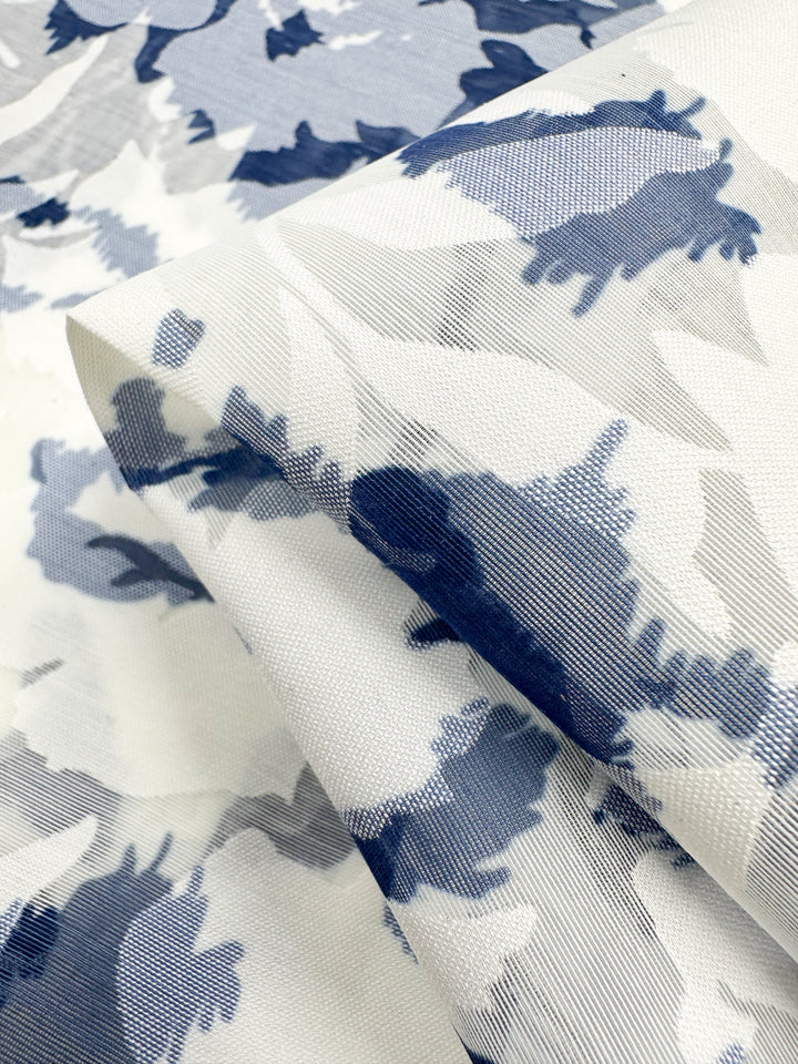 A close-up of Super Cheap Fabrics' Deluxe Print - Blue Flora, a lightweight 150cm polyester fabric with an abstract floral pattern in blue and gray on white. The slightly folded texture highlights its woven details, ideal for dress accessories.