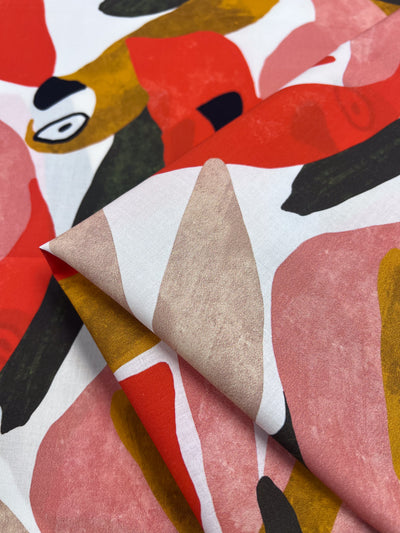 Close-up of Super Cheap Fabrics' Printed Cotton - Tucan, 150cm wide, showcasing bold red, pink, mustard, and green abstract shapes on a white background. The dynamic design highlights vibrant colors and artistic appeal enhanced by natural cotton fibers.