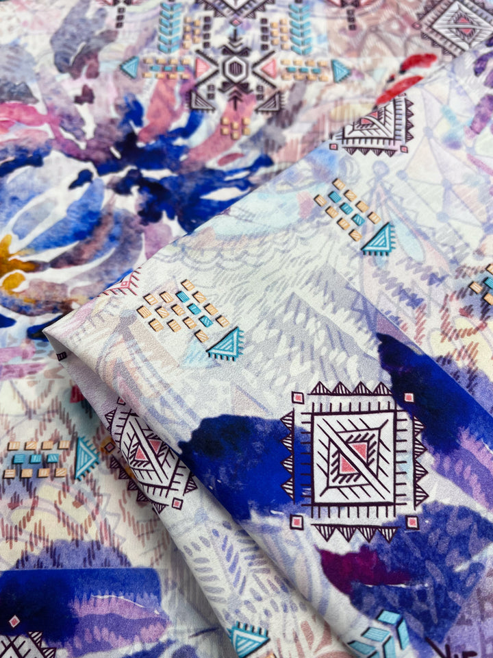 The Printed Cotton - Digital - 150cm by Super Cheap Fabrics is a lightweight fabric featuring a vibrant abstract design with blue, purple, and pink geometric patterns. It blends triangles and squares with watercolor-like floral elements on natural cotton fibers.