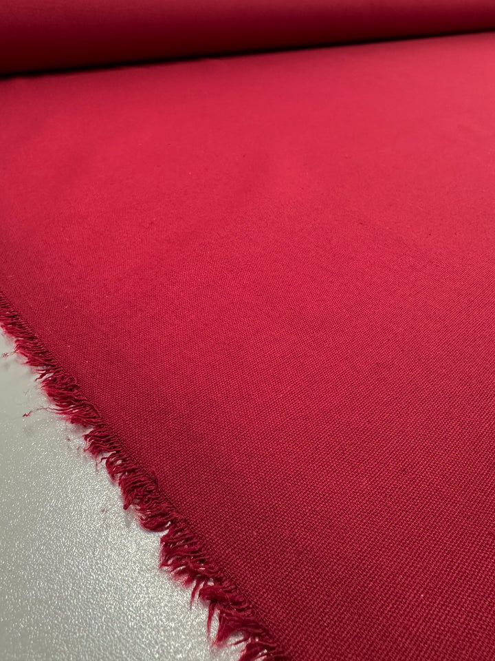 A close-up of a roll of deep red, medium weight Linen Blend - Salsa - 150cm fabric from Super Cheap Fabrics with a slightly textured surface. The fabric edge at the bottom left shows frayed threads, suggesting it is an unfinished cut. The majority of the image showcases the smooth, vibrant red material perfect for elegant table linens.