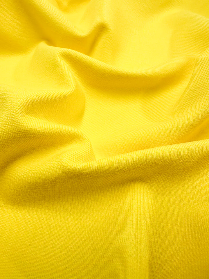 A close-up of Super Cheap Fabrics' Cotton Lycra in Vibrant Yellow (170cm) reveals its smooth texture, gentle folds, and soft, stretchy feel. The well-lit fabric highlights subtle creases and weave patterns.