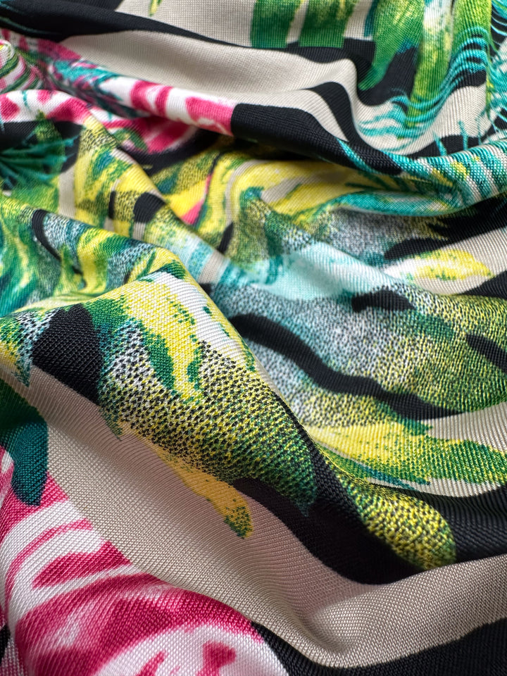 Close-up of Super Cheap Fabrics' Printed Lycra - Jungle Frame, 145cm. The Polyester/Spandex material showcases a tropical design in green, yellow, and pink, with a smooth texture and vibrant patterns gently draped.