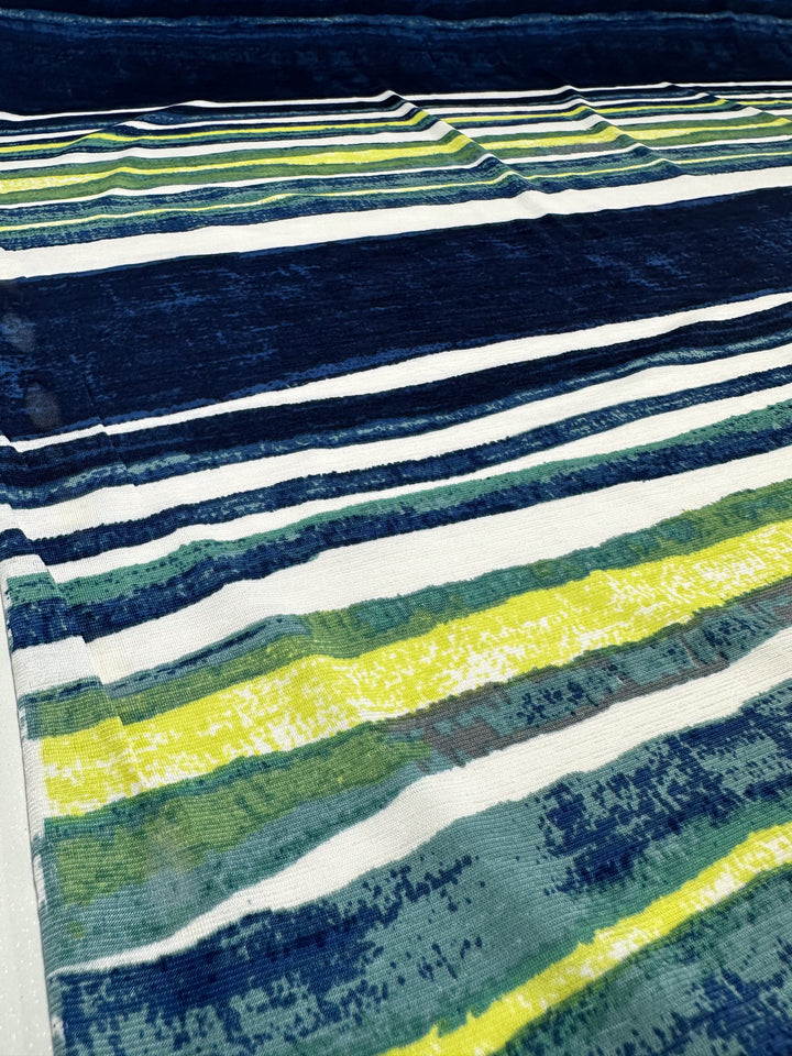 A close-up of Super Cheap Fabrics' Printed Lycra - Ocean Stripe (145cm), featuring horizontal stripes in shades of blue, yellow, and green with a watercolor effect. The Polyester/Spandex blend fabric lays flat, offering a smooth, blended look perfect for summer garments.