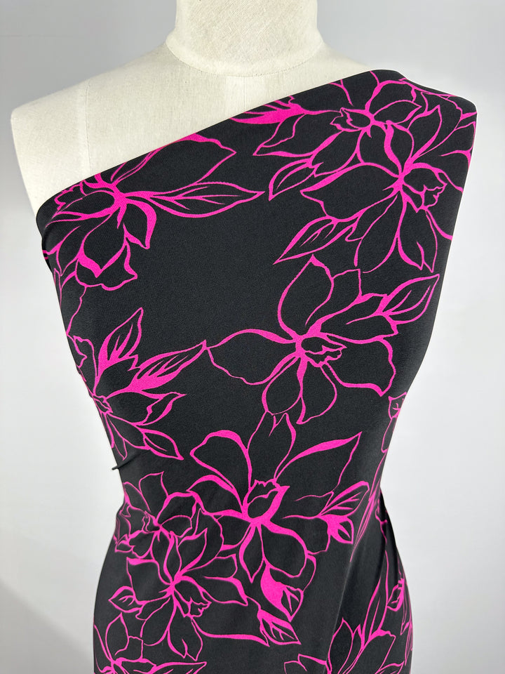 A mannequin features the "Printed Lycra - Devour - 145cm" dress from Super Cheap Fabrics, with a polyester/spandex blend and bold pink floral outlines on a black backdrop, offering comfortable two-way stretch for stylish ease.