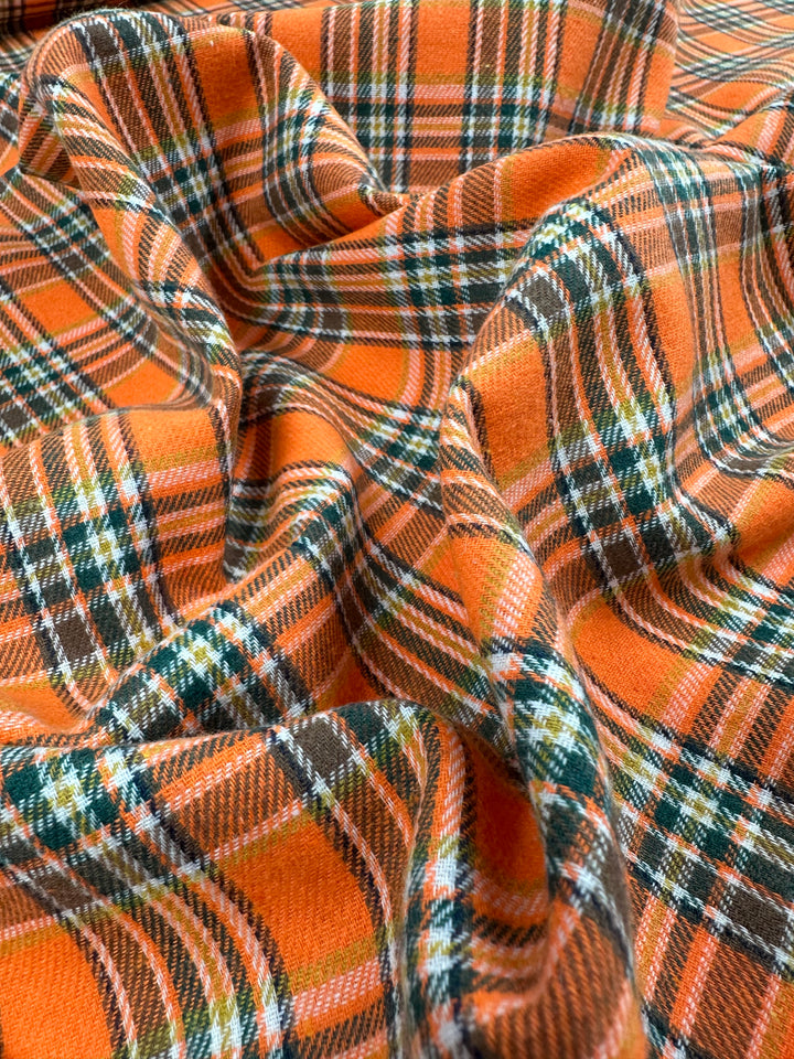 A close-up view of the versatile Suiting - Orange Glen Check from Super Cheap Fabrics. The fabric showcases an intricate combination of orange, green, white, and black lines that form a classic tartan design. The lightweight material drapes softly with slight wrinkles, highlighting its texture and vibrant colors.