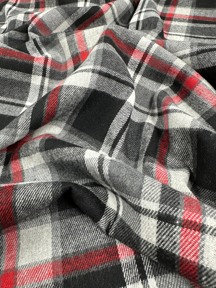 A close-up image of a crumpled fabric featuring the Tartanic suiting from Super Cheap Fabrics, showcasing a plaid pattern in red, black, white, and gray colors. This lightweight fabric appears soft and slightly textured with overlapping lines forming a traditional tartan design—highlighting its versatility for multiple uses. The fabric measures 147cm in width.