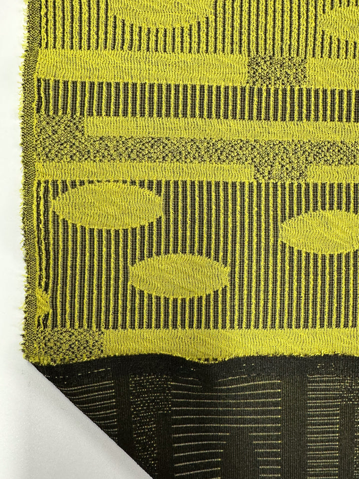 Close-up of Super Cheap Fabrics' Jacquard Knit - Chartreuse (138cm) with a yellow and black pattern showcasing vertical stripes, ovals, and geometric shapes on a white background. The underside reveals its intricate weave and stretch for maximum comfort.