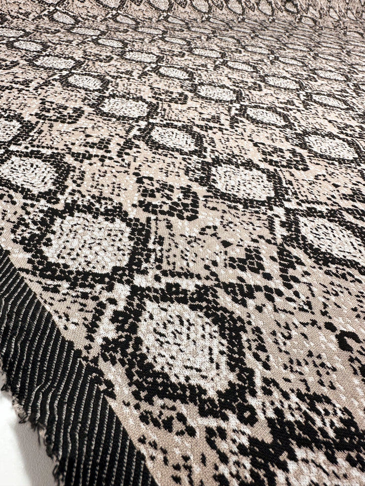 A close-up view of the Jacquard Knit - Mushroom fabric by Super Cheap Fabrics shows a black and beige snakeskin pattern with hexagonal shapes and dotted motifs. This medium-weight, textured knit features a fringed edge, generous stretch, and measures 175cm wide.