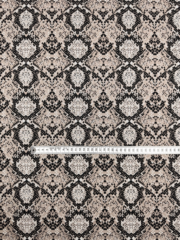 The Jacquard Knit - Mushroom fabric by Super Cheap Fabrics shows intricate black lace patterns on a beige background with symmetrical designs. A measuring tape is placed horizontally across the medium-weight 175cm fabric, displaying scale in both centimeters and inches.