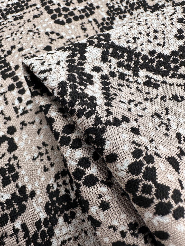 Close-up of Super Cheap Fabrics' Jacquard Knit - Mushroom, featuring a black and white abstract pattern on a beige background. This medium-weight fabric is soft with a generous stretch and design resembling scattered speckles and irregular shapes. Width: 175cm.