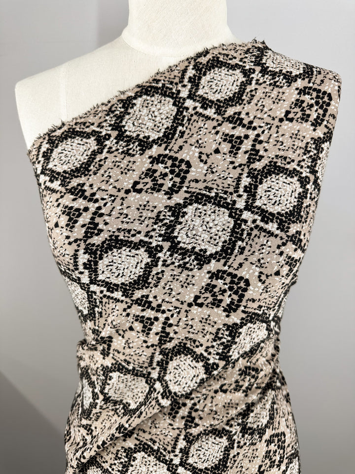 Close-up of a mannequin draped with Super Cheap Fabrics' Jacquard Knit - Mushroom (175cm). This medium-weight fabric features a black and beige snakeskin pattern, known for its generous stretch, styled asymmetrically over one shoulder to highlight its detailed texture and design.