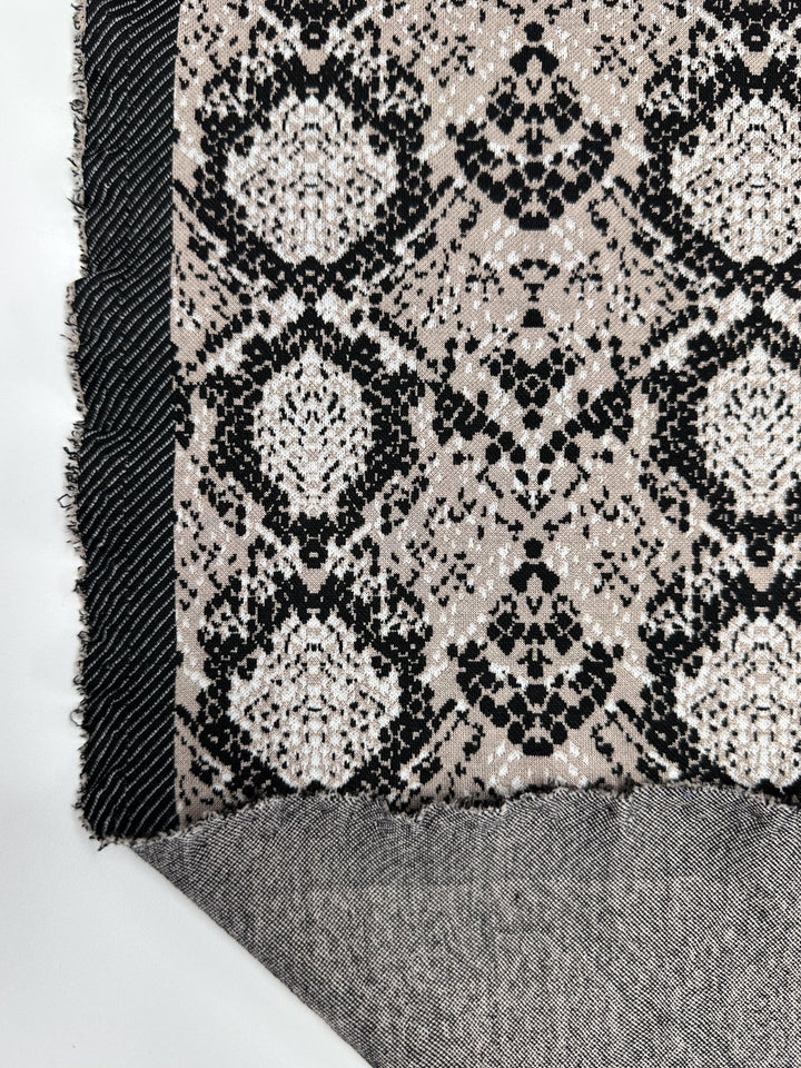 Close-up of Super Cheap Fabrics' Jacquard Knit in Mushroom, 175cm wide, featuring symmetrical black and beige circular and floral designs. A folded edge reveals a simpler black and white woven texture, with a slightly frayed fabric edge.