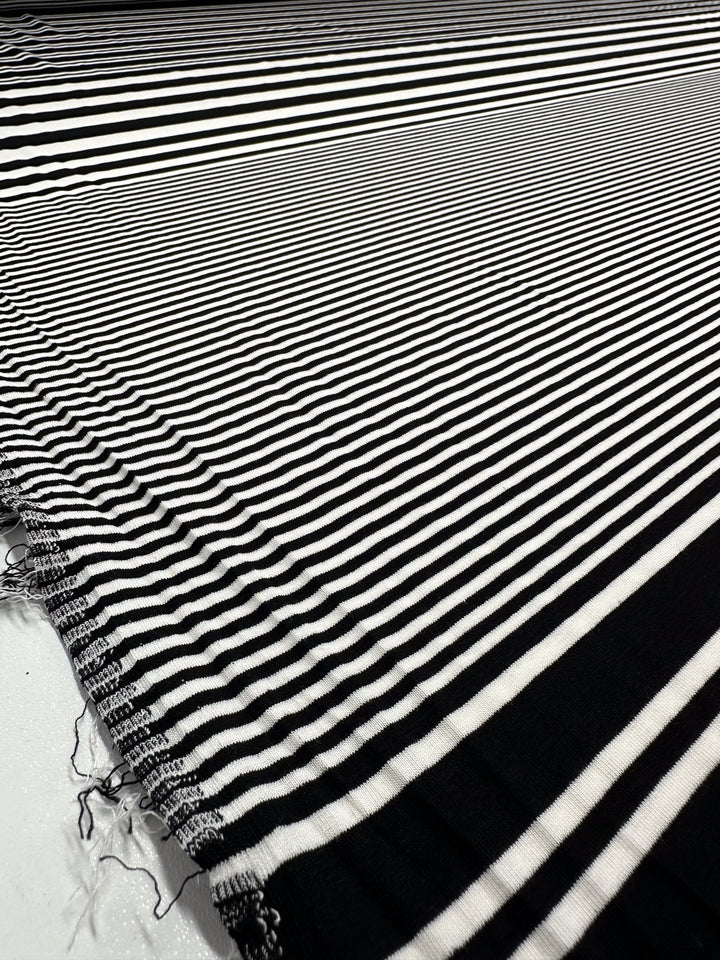 The Viscose Jersey - Paralello from Super Cheap Fabrics unfurls in black-and-white stripes on a flat surface, with alternating lines creating a dynamic width variation. Its slightly frayed edge enhances the fabric's light, soft texture. Width: 160cm.