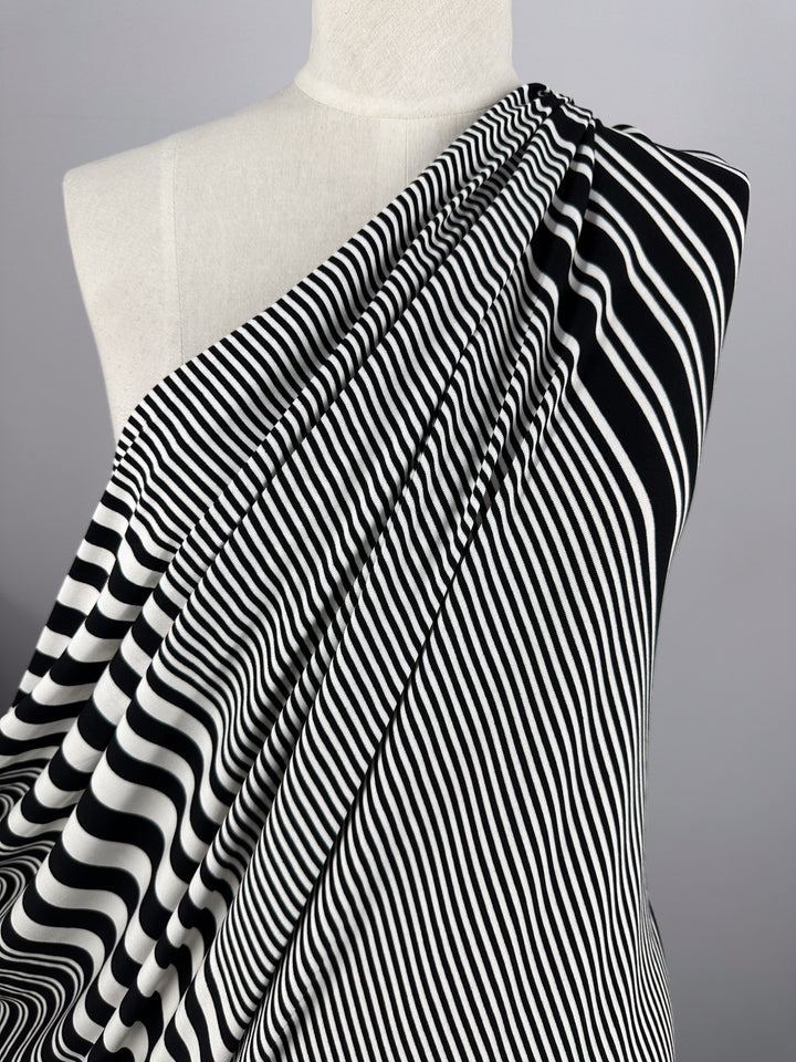 A mannequin's shoulder is draped with Super Cheap Fabrics' Viscose Jersey - Paralello, 160cm. The black and white striped viscose blend features wavy lines that add dynamic movement to the soft, stretchy texture against a plain gray backdrop, highlighting the fabric's flow.