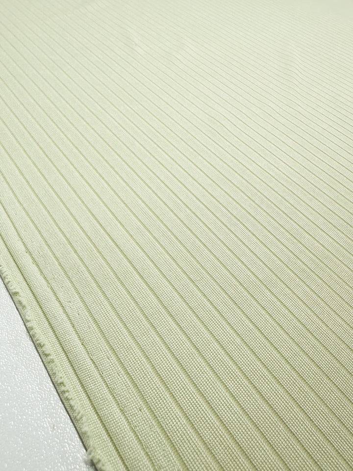 A close-up of Super Cheap Fabrics' Sylvan Green Rib Knit (175cm) displays its light green, medium-weight texture, featuring thin vertical lines. The fabric appears stretchy, comfortable, and subtly reflective with visible edges on one side.