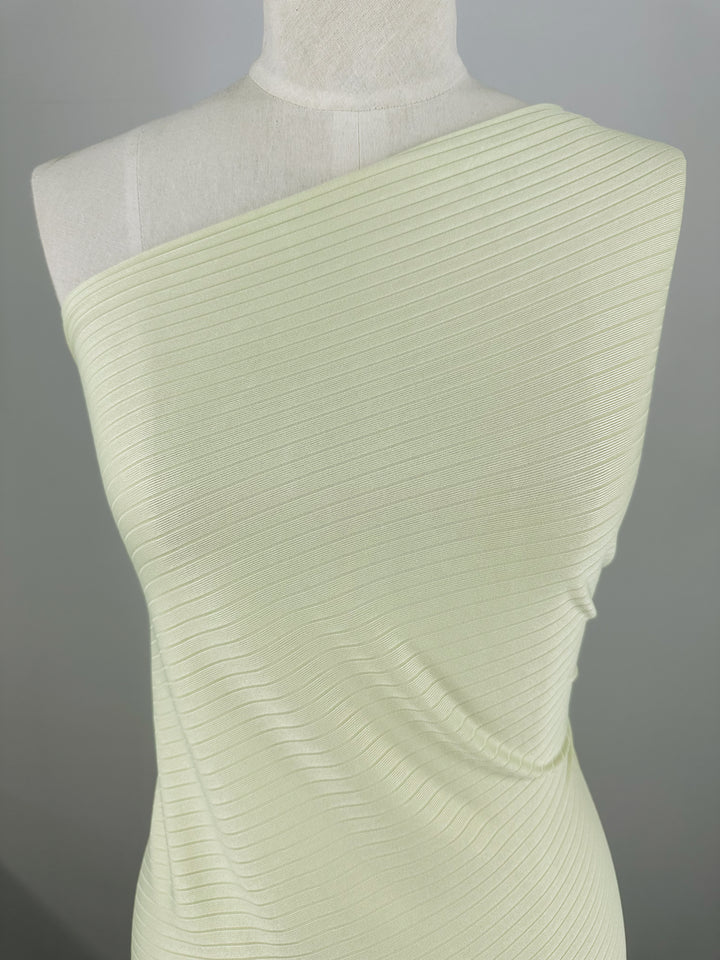 A mannequin displays Rib Knit - Sylvan Green from Super Cheap Fabrics, featuring a ribbed, one-shoulder design. The medium-weight fabric is pale green with diagonal lines, offering a textured look that is both stretchy and comfortable on a neutral background.