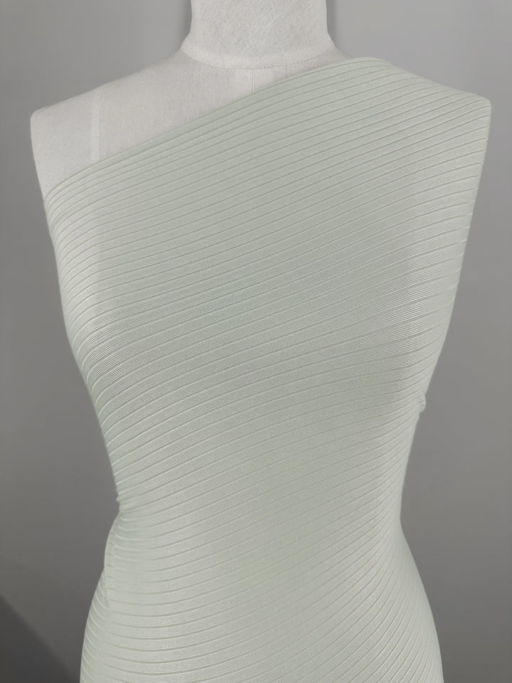 A mannequin showcases the Rib Knit - Murmur garment from Super Cheap Fabrics. It's a one-shoulder design in pale mint green, crafted from medium weight textured rib knit fabric that fits tightly with vertical ribbing. The plain background emphasizes its generous stretch and texture.