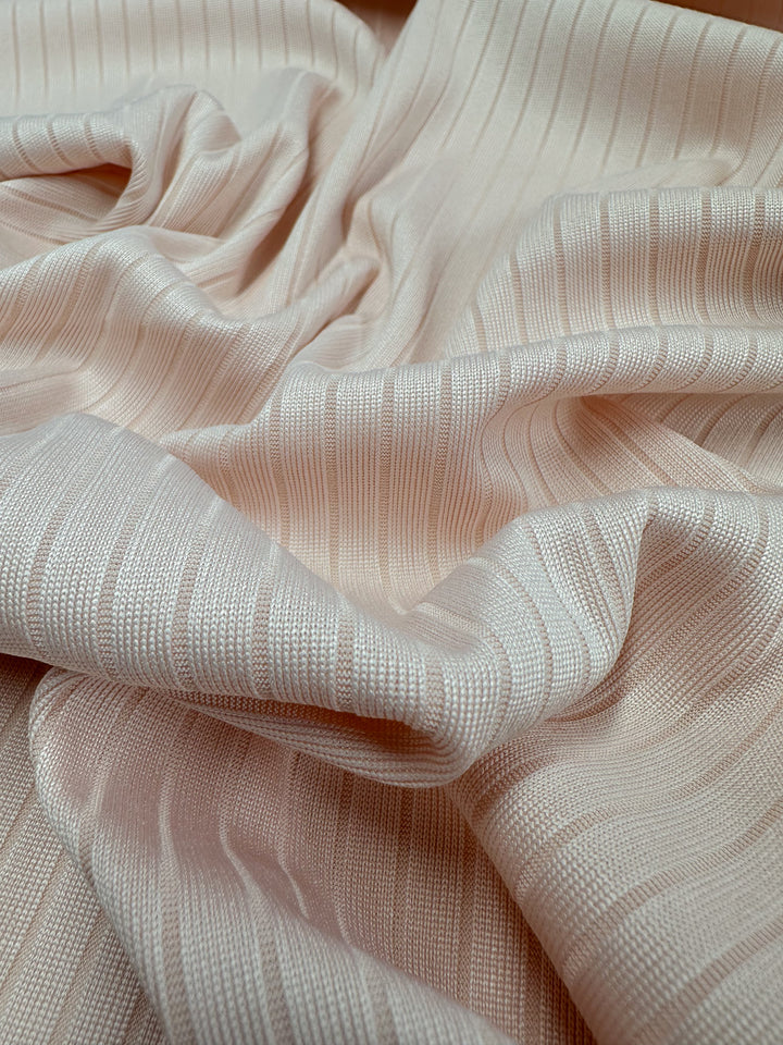 A close-up of a soft Creole Pink rib knit fabric by Super Cheap Fabrics, 168cm wide, showcases its medium weight and textured pattern. The stretchy material is artfully draped and folded, creating gentle waves and shadows while the light enhances its smooth texture and subtle sheen.