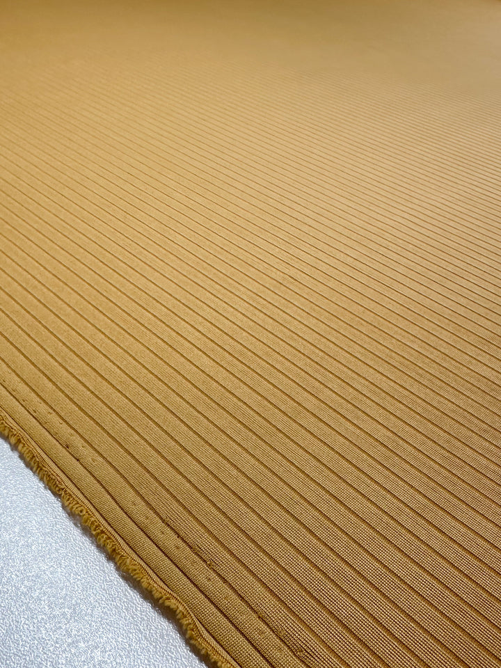 Close-up of Super Cheap Fabrics' Rib Knit in Pale Golden Nugget. This 173cm fabric features a medium weight smooth, textured knit with parallel horizontal lines, a light brown hue, and a slightly frayed border at the bottom.