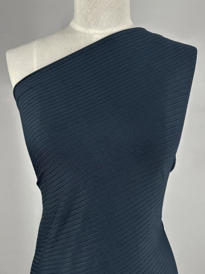 A mannequin dons a Super Cheap Fabrics' Rib Knit - Total Eclipse, 168cm. The dark, medium weight one-shoulder garment showcases horizontal rib patterns with an elegant stretch, set against a plain light gray background to enhance its textured details.